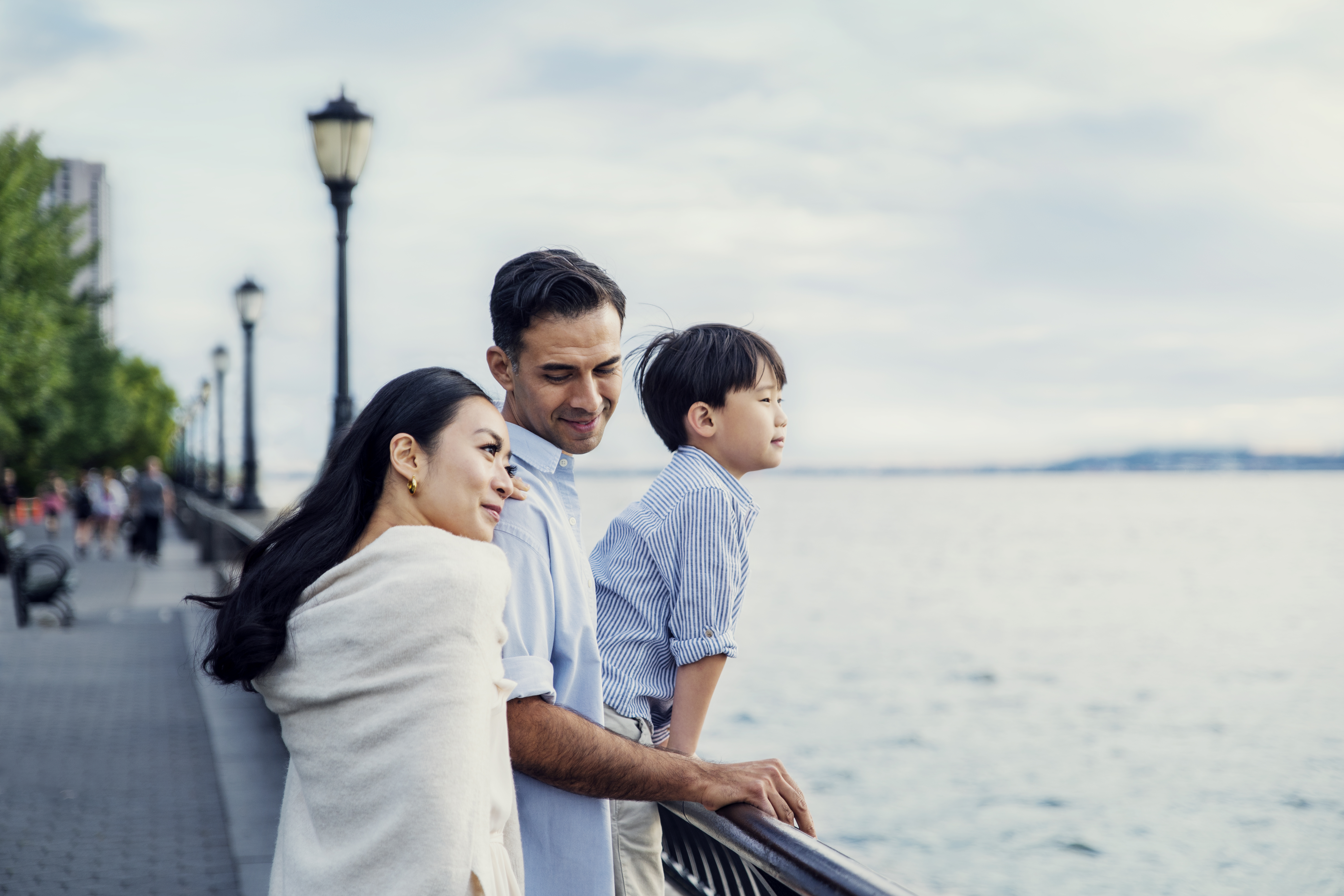 A woman, man, and child sit stand against a railing looking at the Hudson river with green trees behind them.